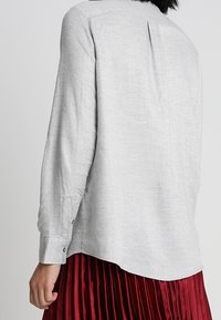 Ljusgrå långärmad skjorta med en mjuk textur, med en plissering bak och knäppningar vid ärmsluten, tillsammans med en plisserad röd kjol.