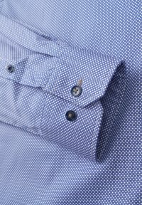 Blaue Bluse mit einem kleinen weißen kreisförmigen Muster, ausgestattet mit einem umgeschlagenen Manschette und zwei dunklen Knöpfen. Die glatte Stoffstruktur ist sichtbar.