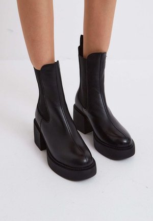 Bottines en cuir noir avec talons épais et semelles compensées portées sur jambes nues sur fond blanc.