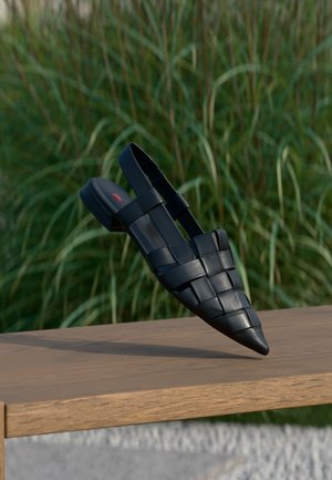 Black woven leather slingback flat shoe balancing on a wooden bench with tall green grass in the background.