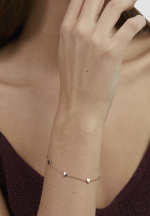 Gold-tone bracelet featuring heart-shaped accents along a delicate chain, displayed on a hand against a maroon textured background.