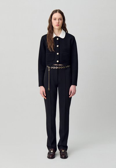 Giacca nera corta con colletto bianco, bottoni argentati, abbinata a pantaloni neri a vita alta e una cintura in catena dorata. Tessuto liscio, vestibilità sartoriale.