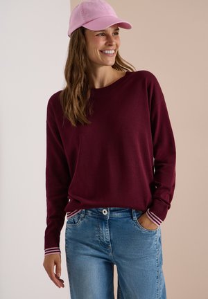 Femme souriante, portant une casquette rose, un pull bordeaux avec des poignets rayés, et un jean bleu, debout avec une main dans la poche.