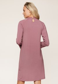 Robe longue à manches longues rose avec une ouverture en forme de goutte dans le dos. Tissu doux, coupe droite, et détails froncés aux manches. Longueur mi-cuisse.