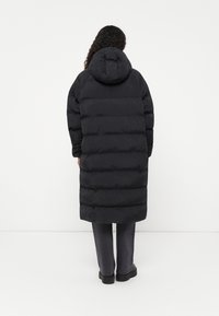 Casaco preto acolchoado com capuz, apresentando um design longo, costuras horizontais e textura suave. Usado com calças cinzentas e sapatos pretos.