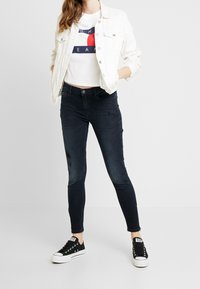 Džínové kalhoty v tmavém odstínu s mírným opotřebením, kombinované s bílým zkráceným tričkem a světle modrou džínovou bundou. Černé tenisky celý vzhled dokončují.