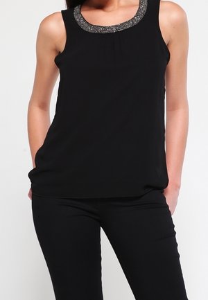 Haut noir sans manches à encolure ronde orné de sequins argentés, porté avec un pantalon noir ajusté.