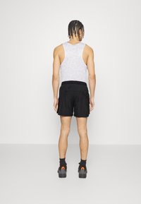 Hombre con el cabello trenzado de pie de espaldas, vistiendo una camiseta sin mangas blanca de malla, pantalones cortos deportivos negros, calcetas negras y zapatillas grises.