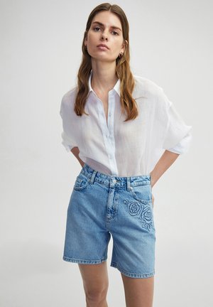 Shorts de mezclilla en azul claro con cintura alta. Presentan un bordado de patrones de rosas en la pierna izquierda y un corte relajado. Combinados con una camisa blanca transparente.