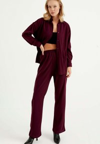 Burgunderfarbenes geripptes Zweiteiler-Outfit mit einem lässig geschnittenen Hemd mit Knopfleiste und Hose mit elastischem Bund. Schwarze High Heels.