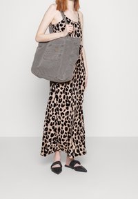 Sac fourre-tout en toile grise avec une poche avant, des bretelles larges et une texture douce, porté avec une robe longue à imprimé léopard et des chaussures noires à enfiler.