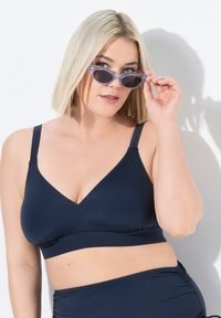 Top da bagno blu navy con design a scollatura a V, tessuto morbido, spalline regolabili e dettaglio arricciato in basso, abbinato a occhiali da sole chiari.