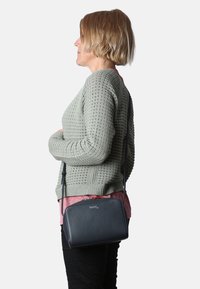 Borsa a tracolla in pelle blu navy con chiusura a zip, texture liscia e un discreto dettaglio con logo, indossata al fianco di una persona contro un maglione pastello.
