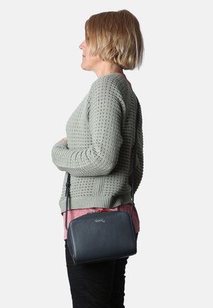 Borsa a tracolla in pelle blu navy con chiusura a zip, texture liscia e un discreto dettaglio con logo, indossata al fianco di una persona contro un maglione pastello.
