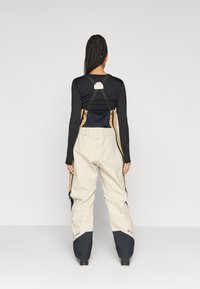 Beige színű sínadrág fekete részletekkel, magas derékkal, állítható pántokkal, oldalsó cipzárakkal és strukturált felülettel.