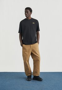 Μαύρο κοντομάνικο μπλουζάκι με μικρό λογότυπο, συνδυασμένο με χακί φαρδιά παντελόνια και μαύρα αθλητικά παπούτσια, σε μονόχρωμο φόντο.