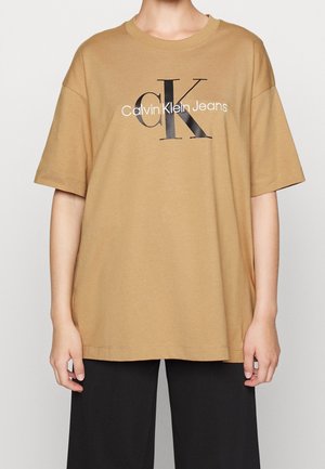Osoba nosząca oversize'ową koszulkę w kolorze wielbłądzim z czarno-białym logo "Calvin Klein Jeans" na piersi, w połączeniu z czarnymi spodniami.