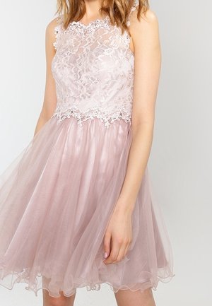 Mouwloze roze kanten jurk met een lijfje met bloemenmotief en een uitlopende tule rok, gedragen door een vrouw met los golvend haar.