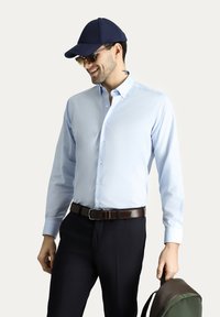 Camicia bluceruleo button-up, maniche lunghe, colletto, abbinata a pantaloni scuri, un cappellino scuro e occhiali da sole, mentre tiene una borsa verde e marrone.