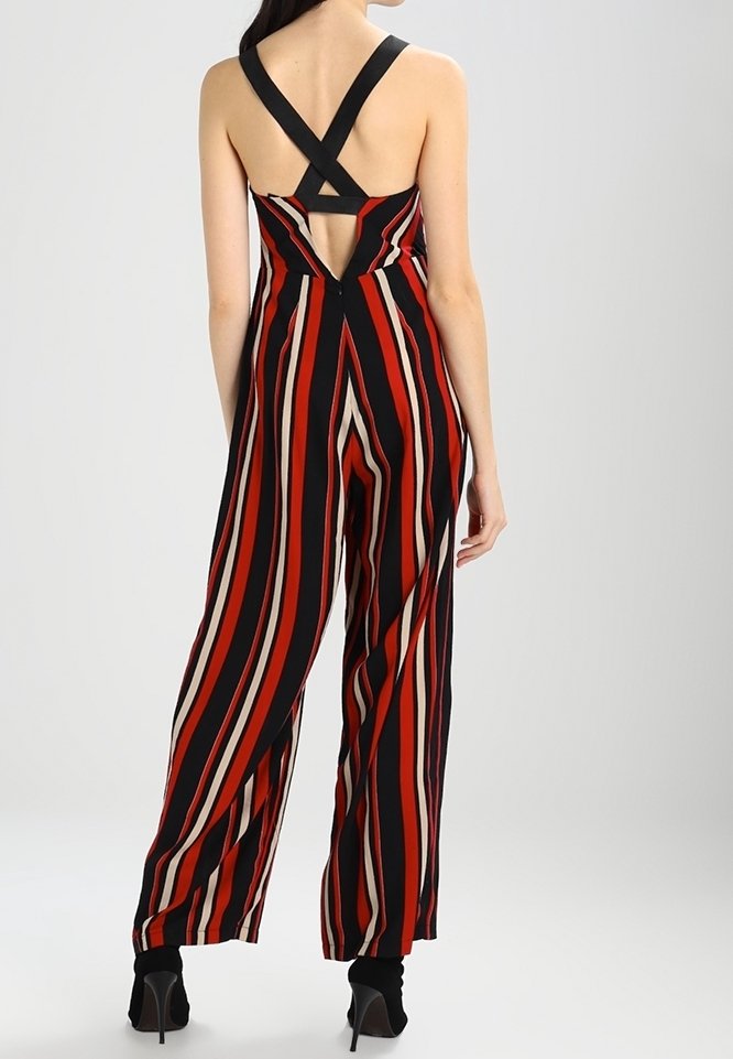 Femme portant une combinaison sans manches à jambes larges avec des rayures verticales rouges, noires et beiges et des bretelles croisées dans le dos, associée à des talons noirs.