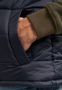 Marineblaues Daunenjacke mit gestepptem Design, ausgestattet mit einer Seitentasche und olivgrünem Rippbündchen auf einem hellbraunen Fleece darunter.