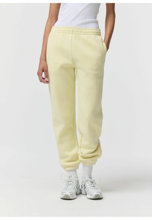 Osoba ubrana w jasne żółte spodnie dresowe, białe skarpetki i białe sportowe trampki, stojąca z rękami w kieszeniach na jednolitym tle.