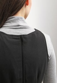 Kvinna sedd bakifrån i en grå polotröja under en ärmlös svart klänning mot en enkel bakgrund.