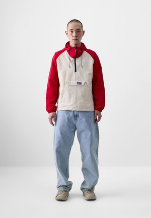 Beige and red zip-up hooded jacket with a front pocket; paired with light blue baggy jeans and beige sneakers. Simple design and materials.
