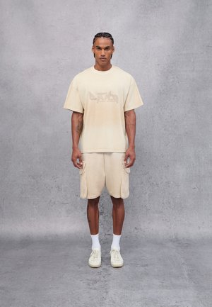 T-shirt oversize beige avec un graphique délavé, shorts beige assortis avec poches latérales et baskets de couleur claire. Texture de tissu lisse.