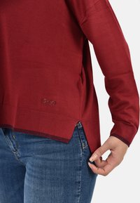 Maglione rosso a maglia con maniche lunghe, fessure laterali e un orlo testurizzato. Etichettato con "LIU JO" vicino all'angolo in basso a sinistra. Indossato con jeans blu.