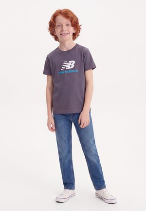 Smilende rødhåret dreng iført en grå New Balance t-shirt, blå jeans og hvide sneakers, der står mod en hvid baggrund.
