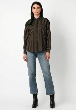 Frau mit langen dunklen Haaren, die ein braun gestreiftes Hemd mit Knöpfen, blaue Jeans und schwarze Knöchelstiefel trägt, steht vor einem einfachen weißen Hintergrund.