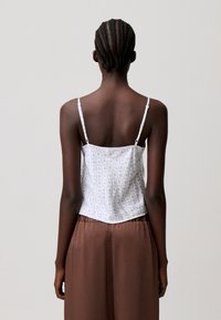 Mujer con el pelo trenzado que lleva una camiseta blanca de tirantes finos con estampado floral y pantalones marrones holgados, de espaldas contra un fondo liso.