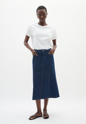 Weißes Kurzarm-T-Shirt, kombiniert mit einem langen, dunkelblauen Jeansrock mit Fronttaschen und kontrastierenden Nähten. Schwarze Flip-Flops.
