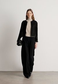 Μαύρο σακάκι με παγιέτες πάνω από μπεζ πουλόβερ, συνδυασμένο με φαρδιές μαύρες παν trousers. Το μοντέλο κρατά μια μικρή μαύρη τσάντα. Λείες, δομημένες υφές.