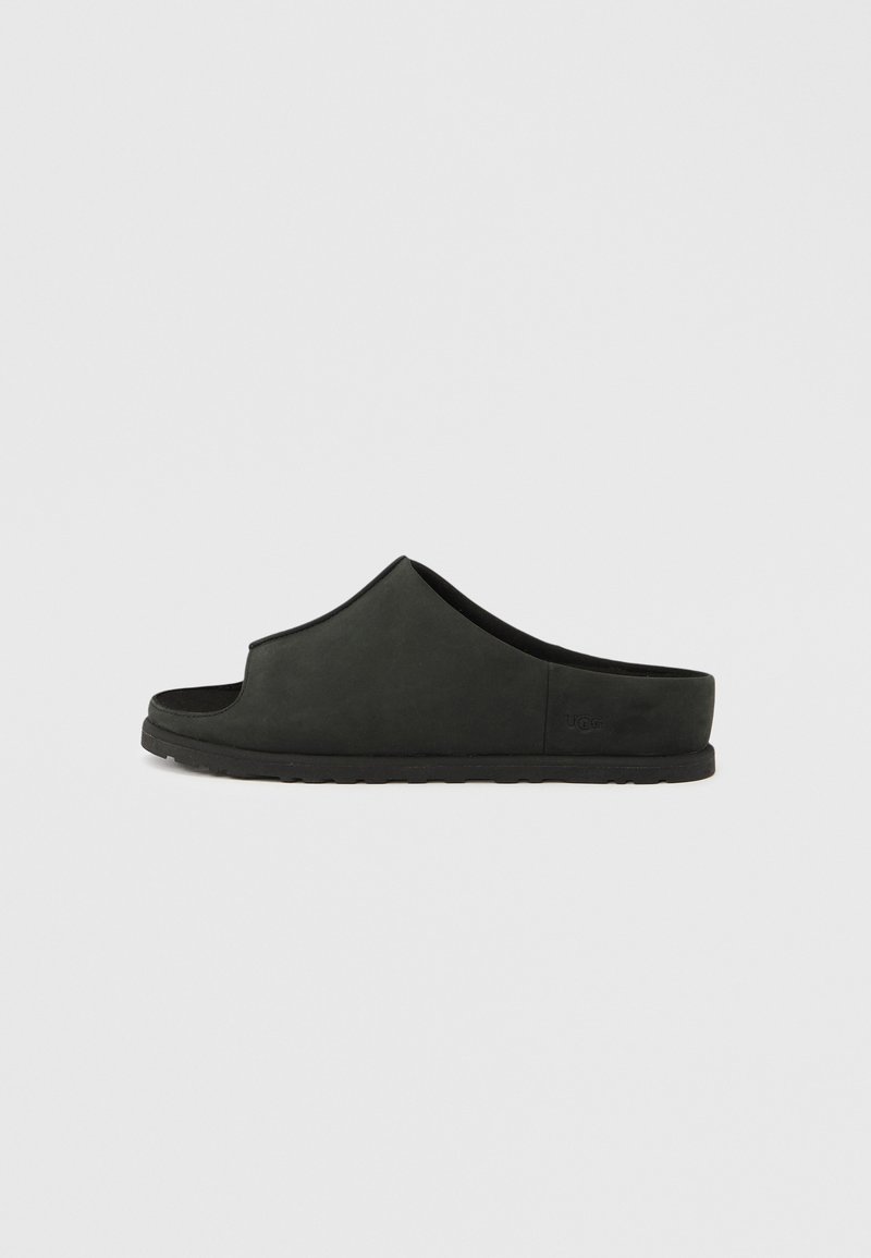 Sandale noire minimaliste à bout ouvert, texture lisse, partie supérieure courbée et semelle plate en caoutchouc. Détail discret de logo embossé sur le côté.