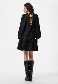 Crna haljina s teksturom, napuhanim rukavima i slojevitom donjim dijelom, s dizajnom leđa s čipkom. Uparena s crnim čizmama s potpeticom do koljena.