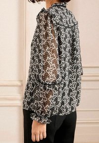 Blusa nera trasparente con motivi floreali e a stelle bianchi, maniche a balze e un bordo nero lungo il collo e l'orlo.