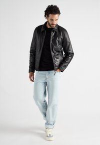 Zwarte leren jas met een gladde textuur, ritsluiting en een aansluitende kraag, gecombineerd met lichtblauwe jeans en witte sneakers.