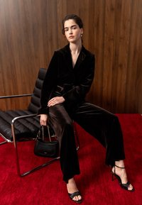 Jeune femme assise sur une chaise en cuir noir portant un costume en velours foncé et des talons noirs, tenant un petit sac à main noir sur un tapis rouge vif.