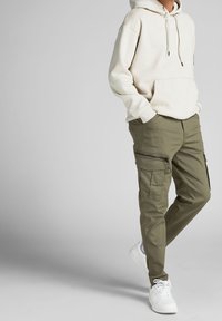 Person, der einen beigen Hoodie, olivegrüne Cargo-Hosen mit Seitentaschen und weiße Sneakers trägt, steht mit den Händen in den Taschen vor einem grauen Hintergrund.