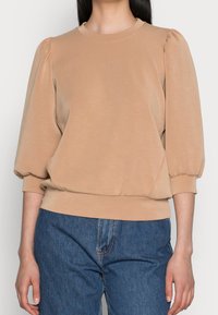 Sudadera beige con mangas abullonadas, escote redondo y dobladillo acanalado. Confeccionada en una tela suave y ligera. Combinada con jeans de mezclilla azules.