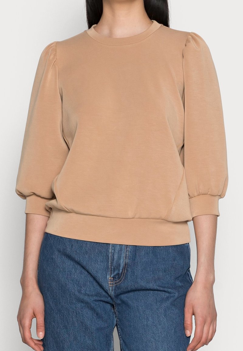 Sudadera beige con mangas abullonadas, escote redondo y dobladillo acanalado. Confeccionada en una tela suave y ligera. Combinada con jeans de mezclilla azules.