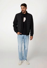 Giacca imbottita nera con collo alto, chiusura con zip e maniche lunghe, abbinata a un maglione scuro, jeans azzurri e scarpe da ginnastica bianche.