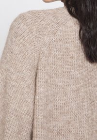 Kvinna med långt mörkt hår som bär en mjuk, stickad beige tröja, visad från axeln och sidan av nacken.