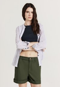 Camicia a righe bianche e blu con bottoni, indossata sopra un crop top nero a coste, abbinata a pantaloni corti verde oliva con risvolti. Texture liscia in tutto l'outfit.