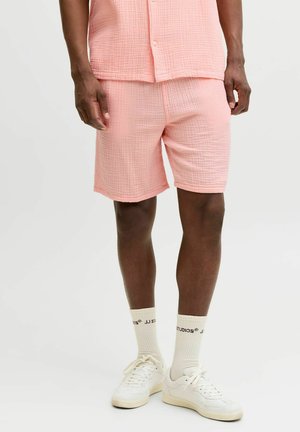 Muž nosiaci svetloružové textúrované šortky a zodpovedajúcu košeľu, biele ponožky s čiernym textom a biele tenisky na jednoduchom pozadí.