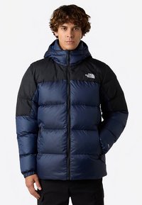 Navy blue puffer jacket with black shoulders, high collar, zip closure, and elastic cuffs. Features a prominent logo on the chest.