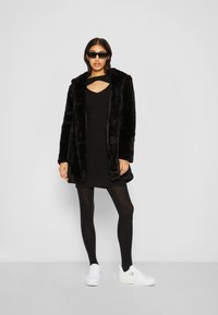 Cappotto in pelliccia sintetica nera con collo a scialle, indossato sopra un vestito nero, collant neri e sneakers bianche. Gli occhiali da sole completano il look.