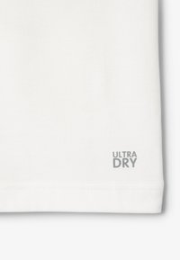 Tela de camiseta blanca que absorbe la humedad con costuras planas; presenta un pequeño texto gris "ULTRA DRY" cerca del dobladillo. Textura suave.